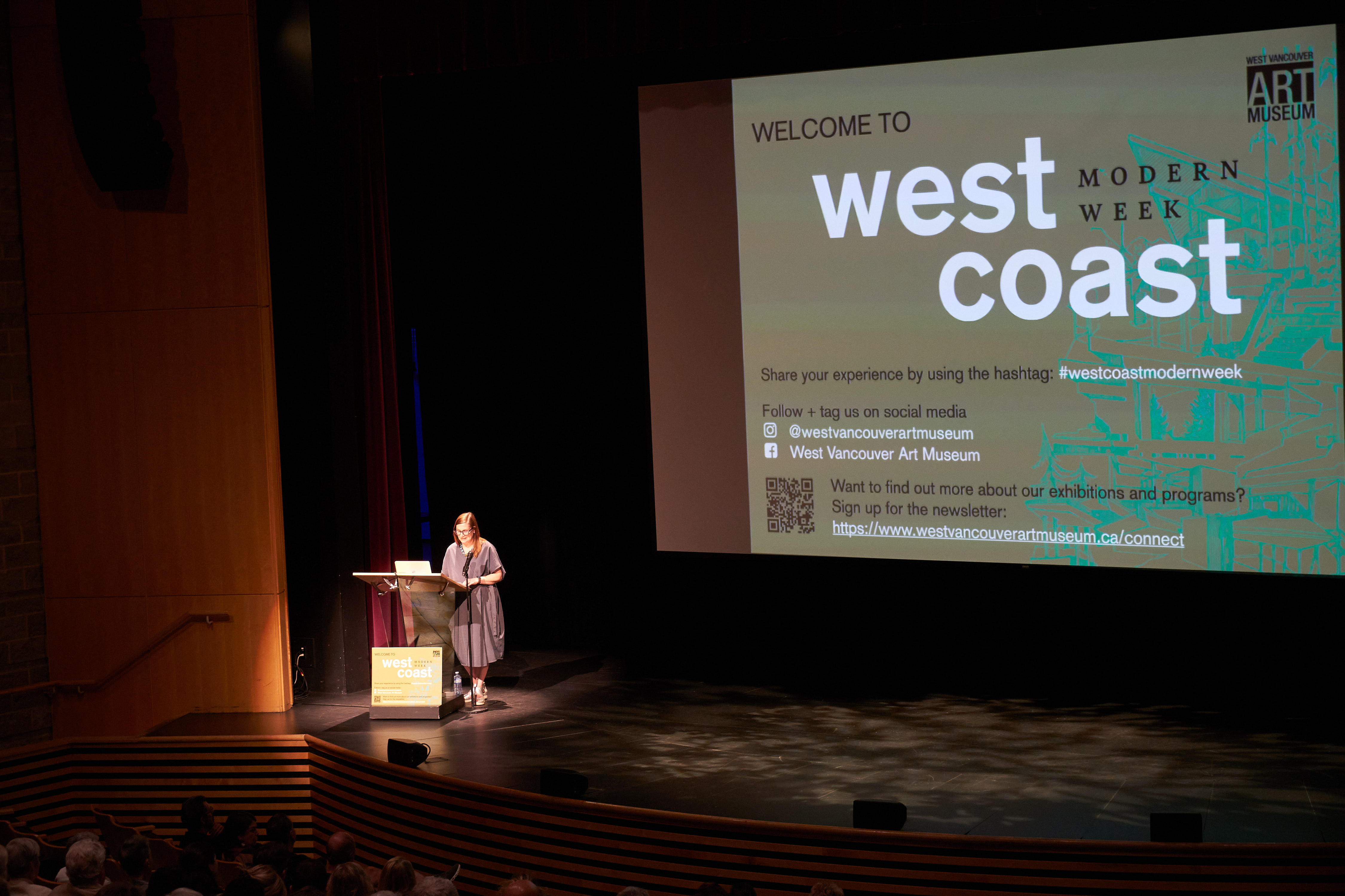 A woman on stage in a large theatre giving a presentation. On the screen behind her is a slide that says "Welcome to West Coast Modern Week" with details about where to find more information on social media and the art museum website.