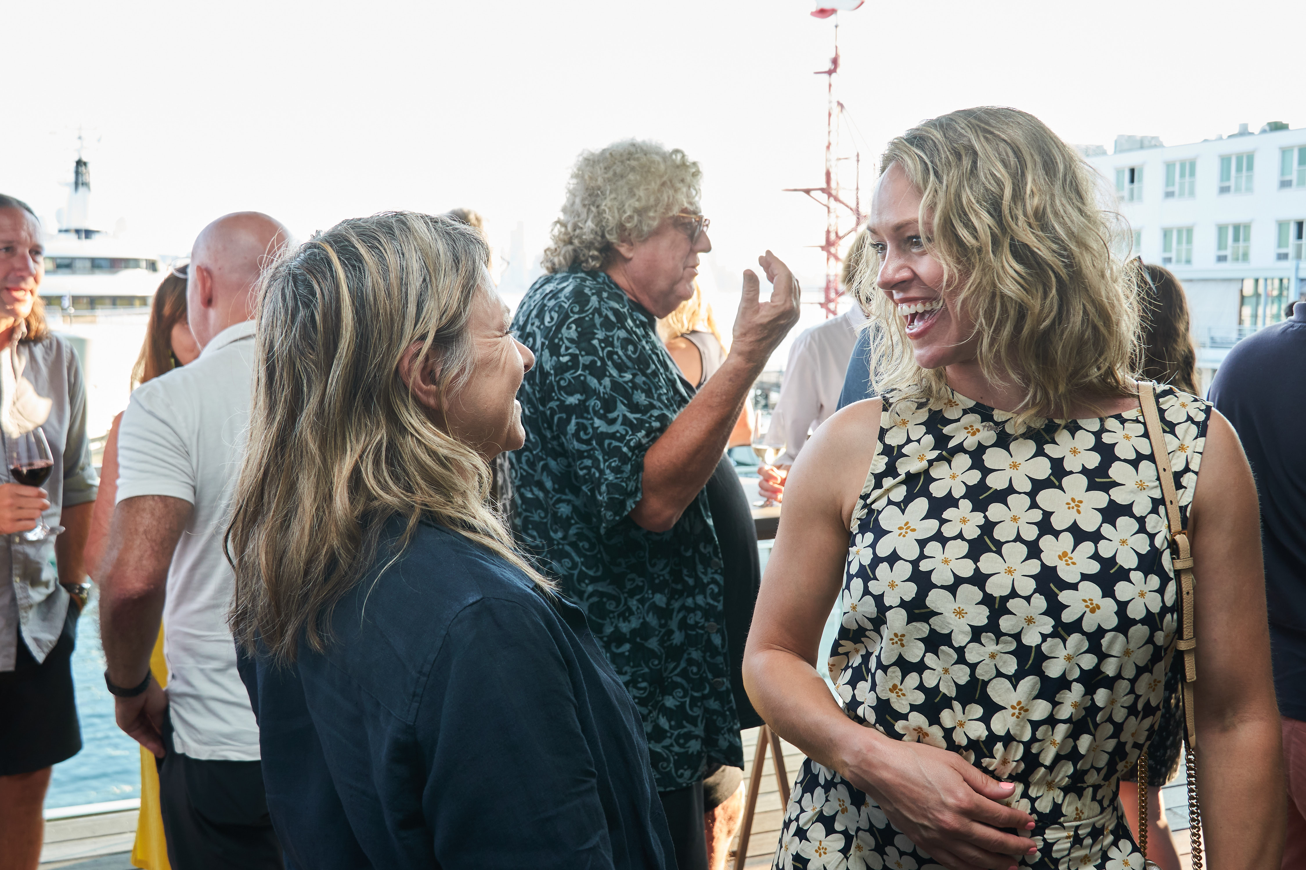 Two women talking and smiling at an outdoor event where people are chatting and drinking wine.