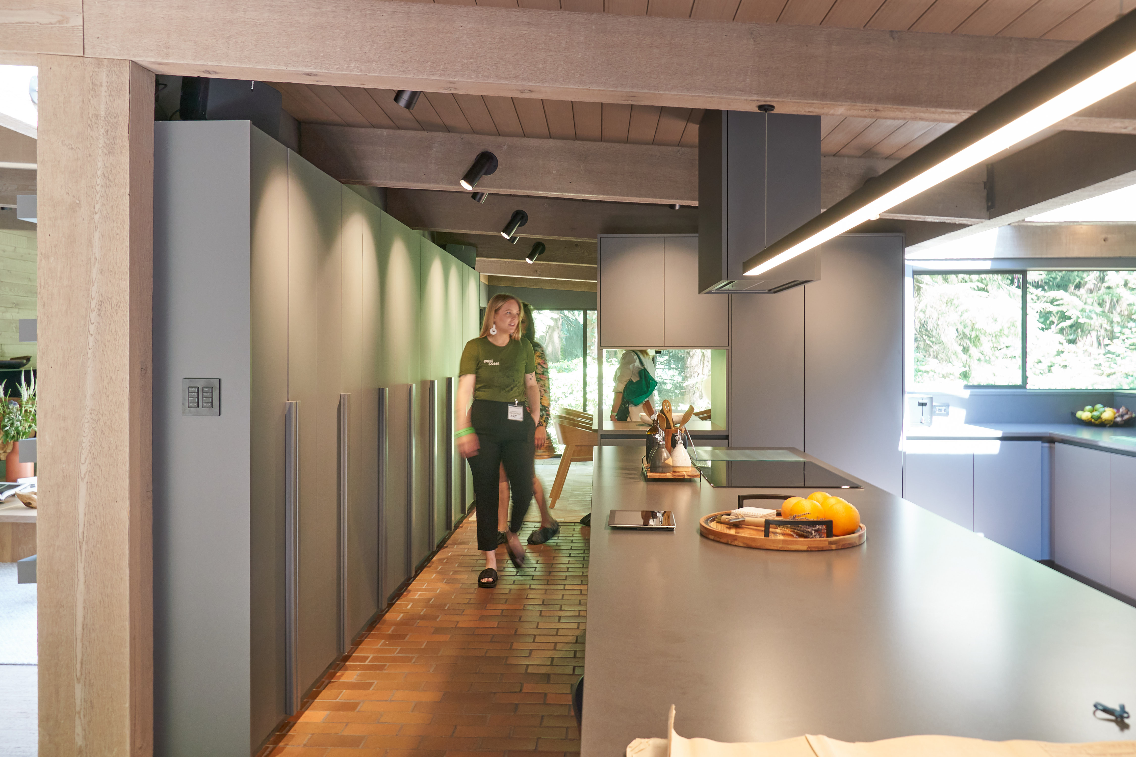 A few people walking through a large kitchen with brick floors, grey cabinets and countertops, and an island.