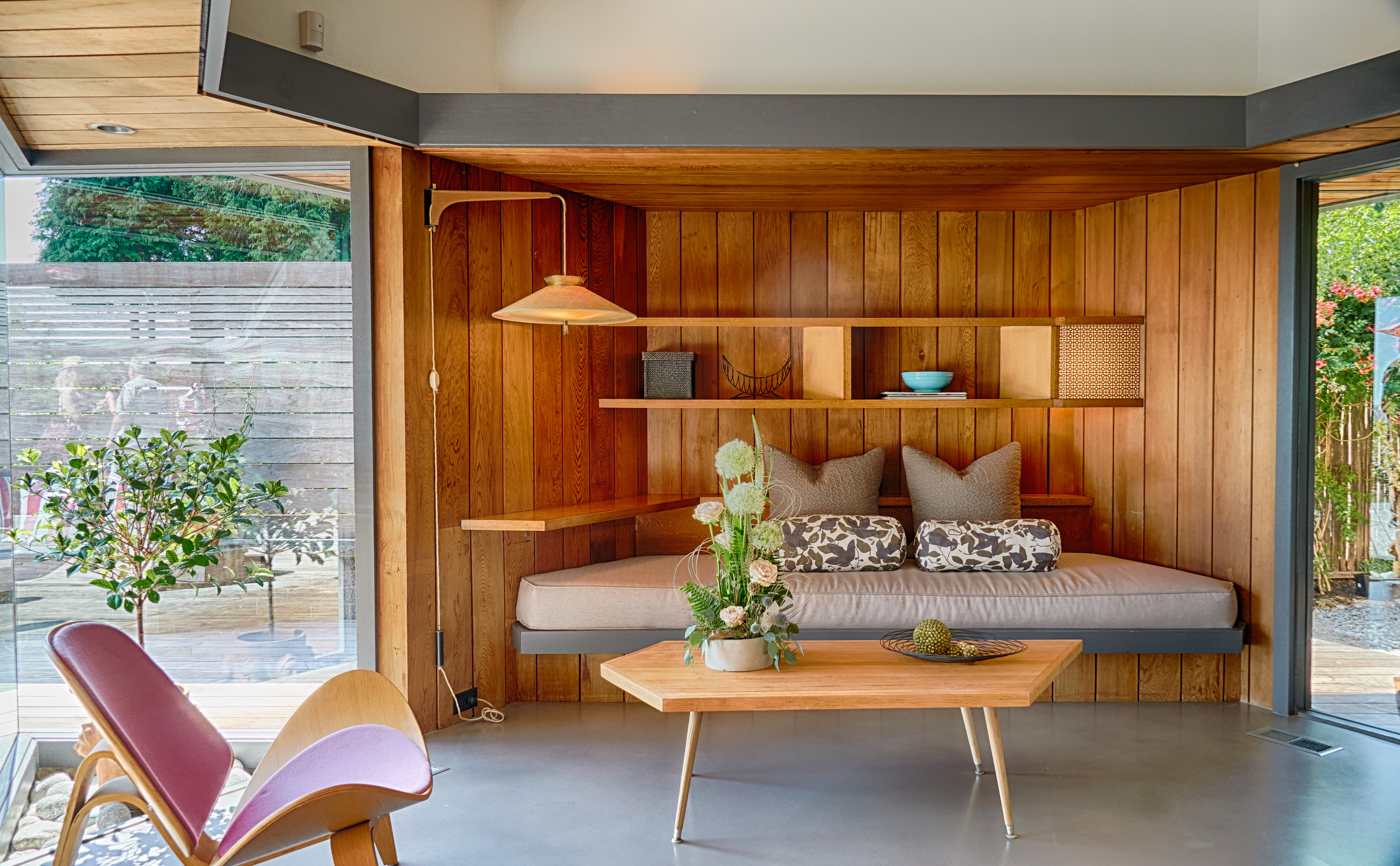 An enclosed outdoor space with glass walls on each side, a wooden alcove with a shelf-style bench, a coffee table and chair.